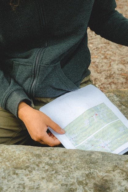 A close-up shot of a hiker's hands adjusting a map and compass on the Appalachian Trail. The trail is visible in the background, winding through a dense forest with dappled sunlight filtering through the trees. The hiker is using the tools to navigate and plan their route.