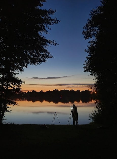 A photographer's setup with a tripod-mounted camera capturing the vibrant autumn foliage reflected in a calm lake during the golden hour in New England, with camera gear visible.