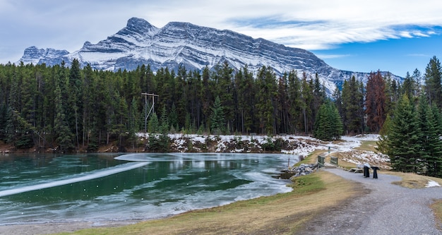 A breathtaking panoramic view of Banff National Park in Alberta, Canada, showcasing turquoise lakes, snow-capped mountains, and dense forests. A solo female traveler is standing on a viewpoint, taking in the scenery with a sense of awe and wonder.