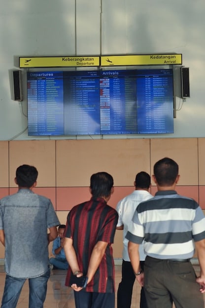 A crowded airport gate with passengers looking at a flight information display board showing multiple delayed and cancelled flights.