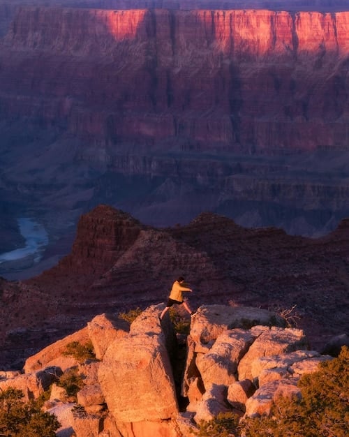 A scenic view of Mesa Arch at sunrise in Canyonlands National Park, Utah, with a person standing on the cliff edge looking out at the vast canyon below.